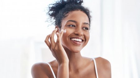 Smiling woman demonstrating how to apply cream to her cheek as part of her clear skin routine.